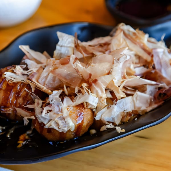 Hikari Ramen's takoyaki, topped with bonito flakes and drizzled with sauce, on a black plate.