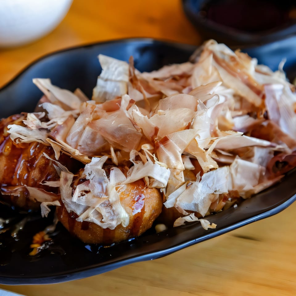 Hikari Ramen's takoyaki, topped with bonito flakes and drizzled with sauce, on a black plate.
